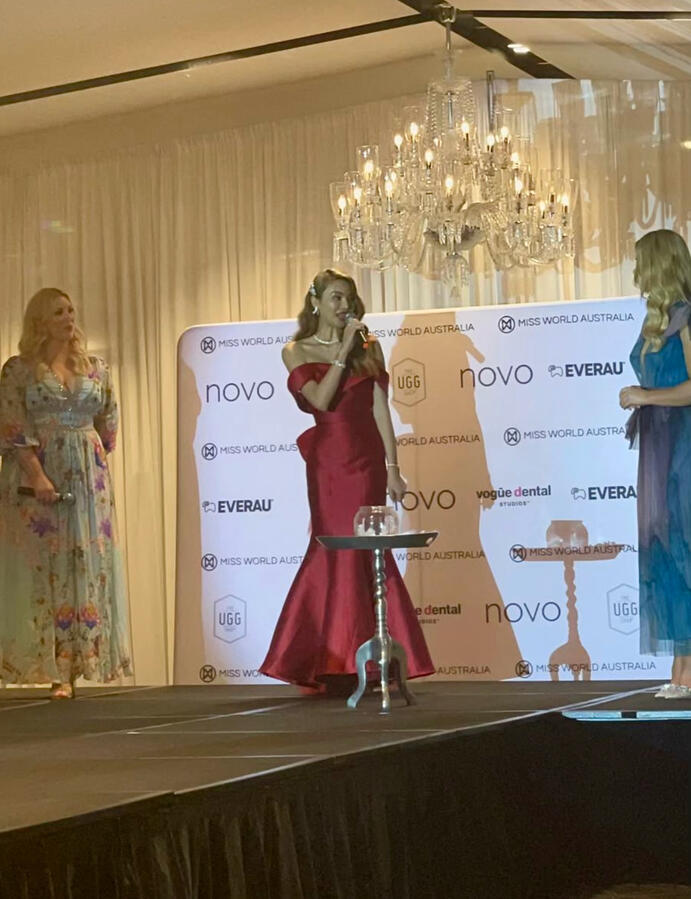 Lenny Cheng in a red gown speaking onstage during a Miss World Australia event in Melbourne, with judges and branding in the background.