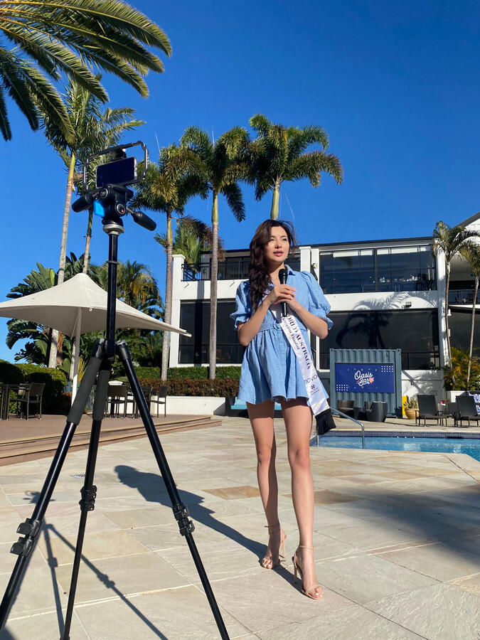 Lenny Cheng wearing a blue dress and Miss World sash, speaking poolside to press during a media engagement at Queensland University.