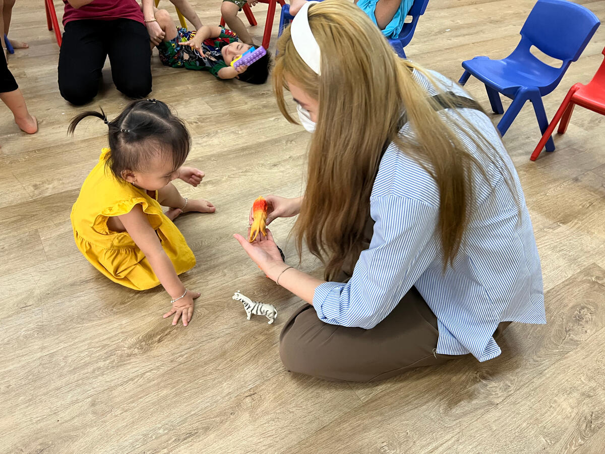 Lenny Cheng kneeling on the floor at a Vietnamese children’s centre, interacting with a young child using animal toys during a volunteer session.